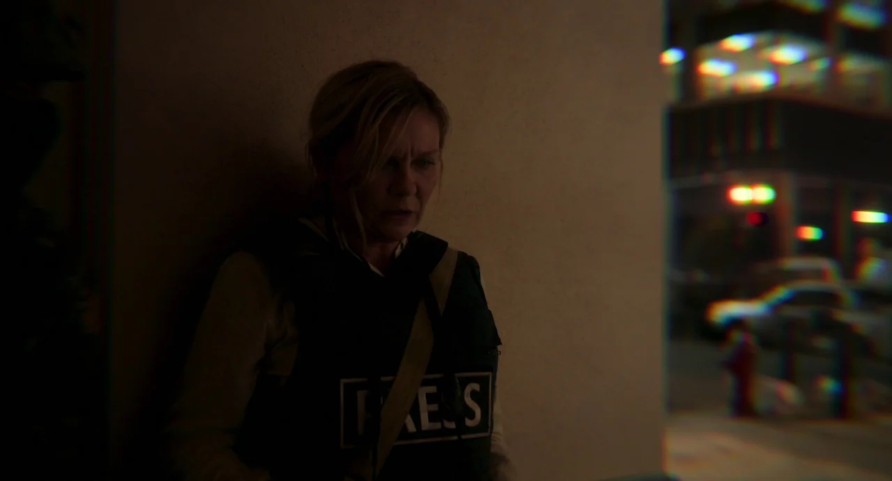 Civil War (2024) movie still: medium — A woman stands in the shadows of an exterior wall at night. Distant colored lights blur in the background…