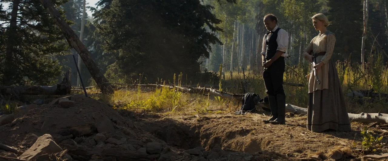 Damsel (2018) movie still: fullbody — A clearing in a dense woodland features an freshly dug dirt grave. A man stands with head bowed next to a…