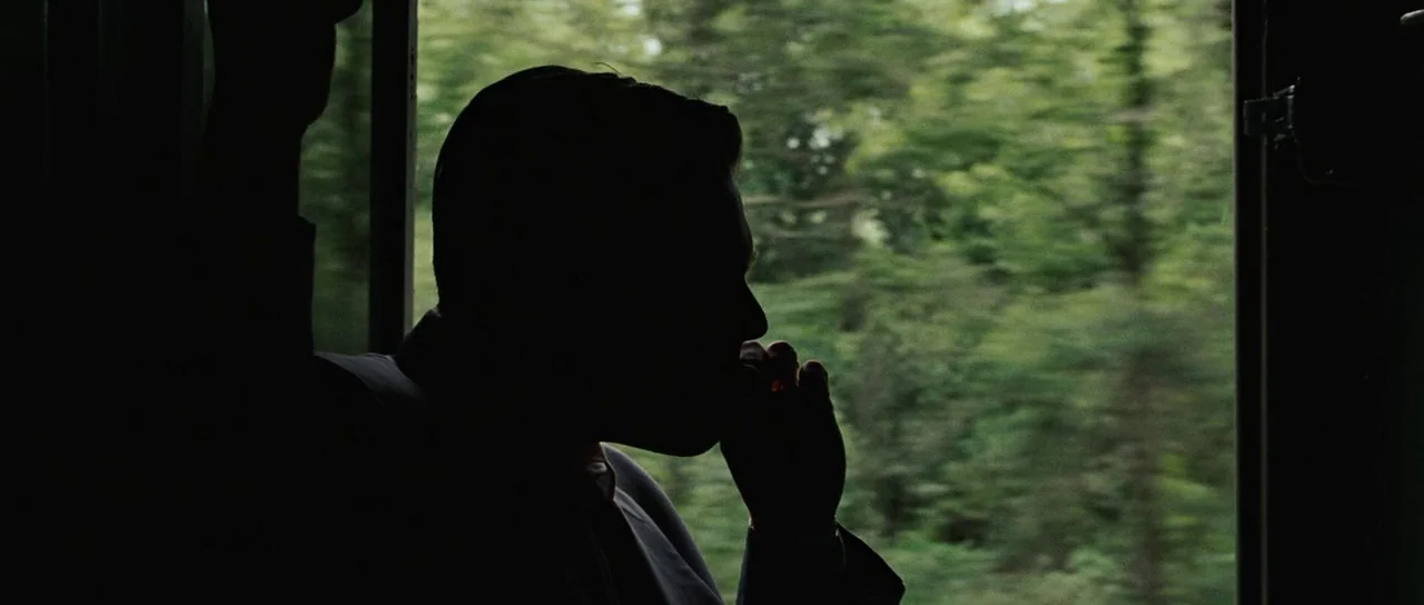 Revolutionary Road (2008) movie still: medium — A man sits inside a train car with a window showing passing green trees. The man is silhouetted…