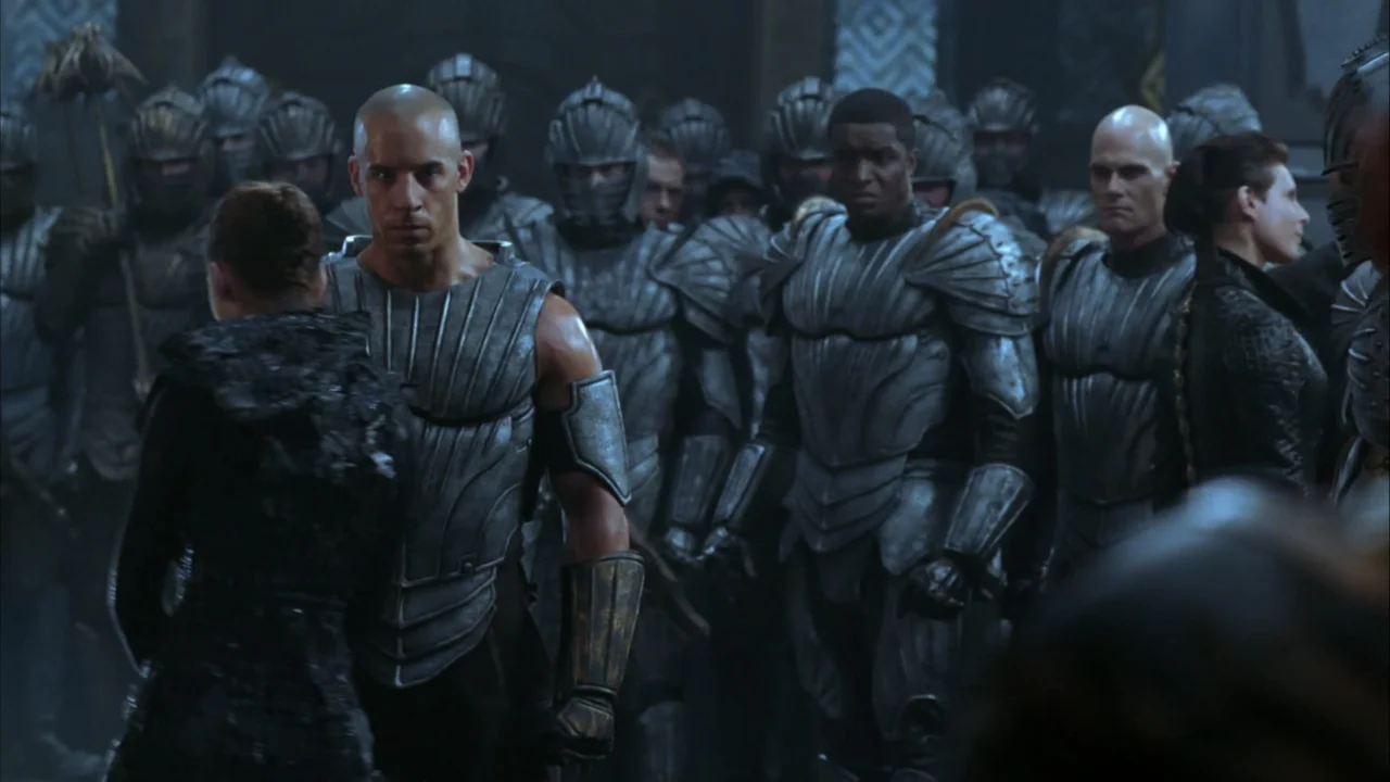 The Chronicles of Riddick (2004) movie still: medium — A stone hall filled with a crowd of soldiers wearing intricate metallic plate armor. A bald…