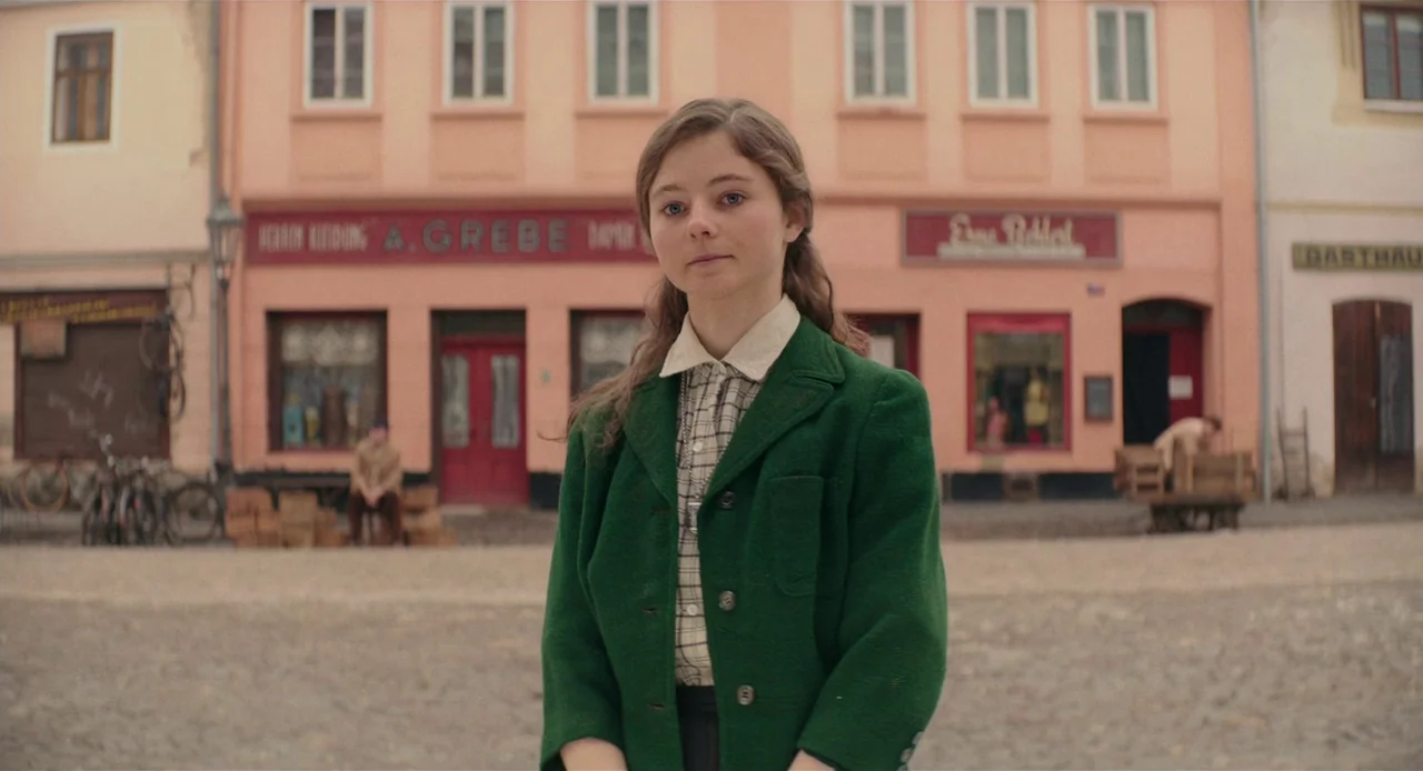 Jojo Rabbit (2019) movie still: medium — A quiet cobblestone town square is lined with pink and beige multi-story commercial buildings. A young girl…
