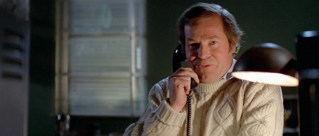 The Fog (1980) movie still: medium — A dimly lit room features a man sitting near a bright desk lamp. The man is holding a black telephone receiver…