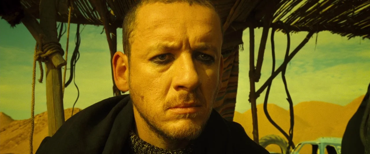 Micmacs (2009) movie still: closeup — A man stands under a shaded structure within a vast, arid desert landscape. The subject faces the camera while…