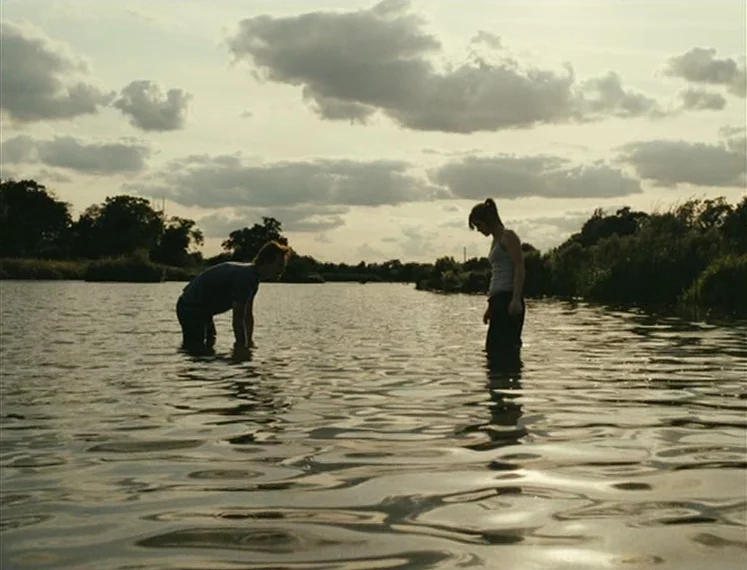 Fish Tank (2009) movie still: wide — A wide, calm river reflects the overcast sky during late afternoon. A man and woman stand knee-deep in the…
