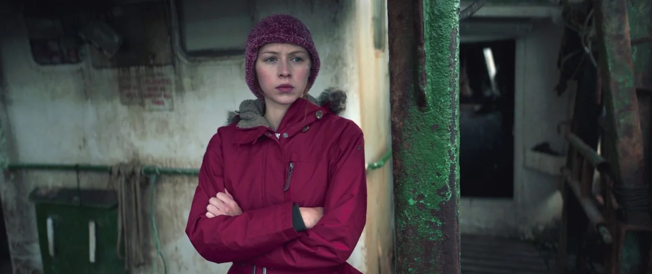 Sea Fever (2019) movie still: medium — A woman stands on the weathered deck of a metallic fishing vessel. She keeps her arms folded across her chest…
