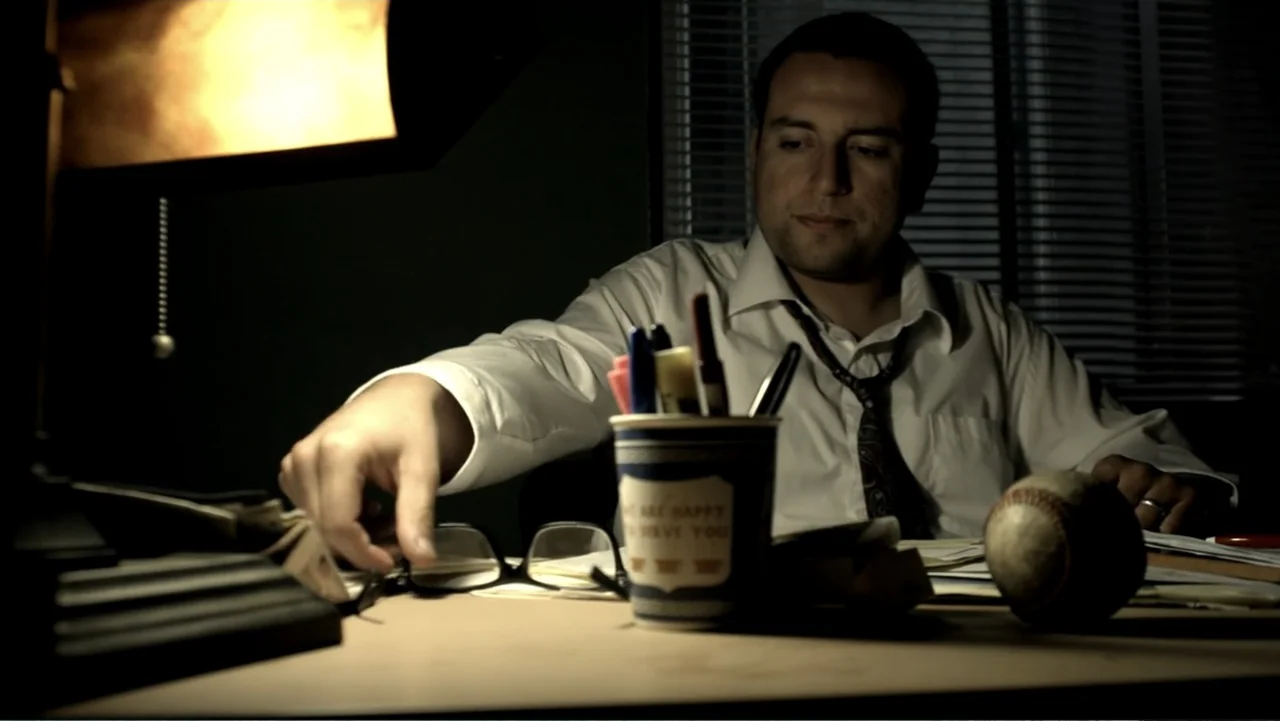 Abandoned Dead (2015) movie still: medium — A dark office environment features a cluttered desk illuminated by a nearby lamp. A man in a dress shirt…