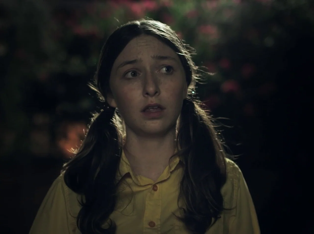Girl Asleep (2015) movie still: closeup — A girl is positioned outdoors against a dark, blurry background of foliage. The subject stands facing the…
