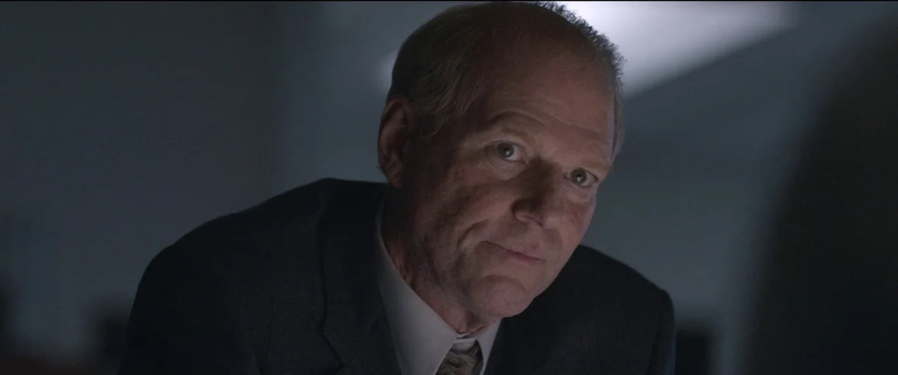 The Good Nurse (2022) movie still: closeup — An older man sits in a dark office space with soft ambient light. He looks directly at the viewer with…