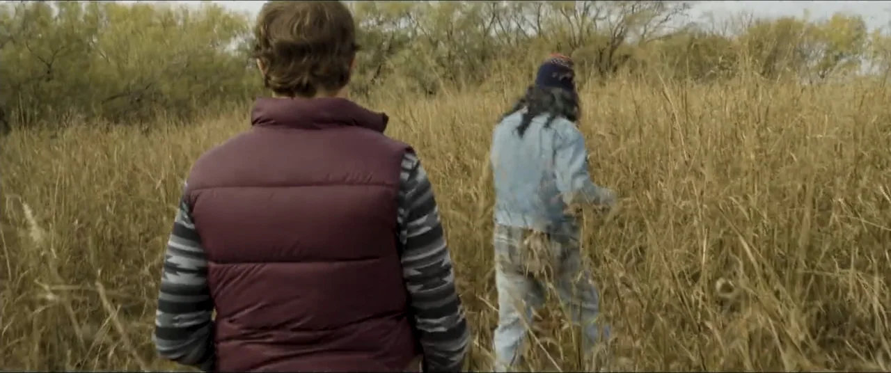 Amityville Cult (2021) movie still: medium — A field filled with tall yellowing dry grass stretches towards a treeline backdrop. Two people move…