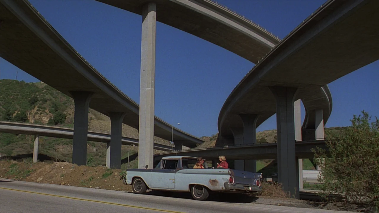 Paris, Texas (1984) movie still: wide — A vintage pickup truck sits parked on the shoulder beneath a towering concrete highway interchange. Two…