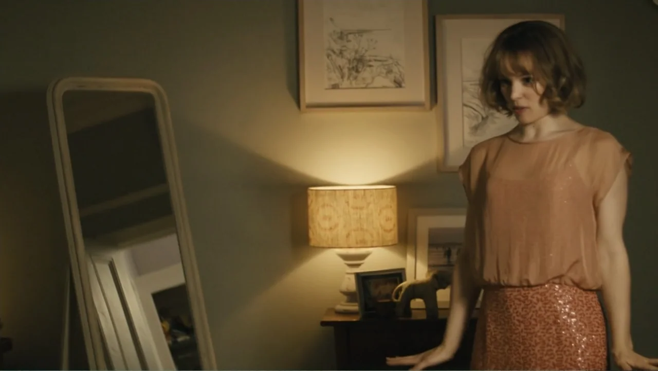 About Time (2013) movie still: medium — A dimly lit bedroom features a floor mirror beside a wooden nightstand with a lamp. A woman wearing a…