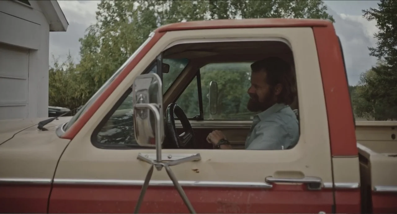 Cowboys (2014) movie still: medium — A vintage two-toned pickup truck sits parked near a white building with trees behind. A man with a beard sits…