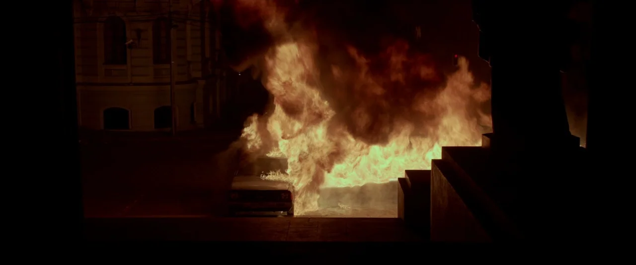 Ema (2019) movie still: wide — A dark city street at night shows a car engulfed in bright orange flames. Architectural features of nearby buildings…