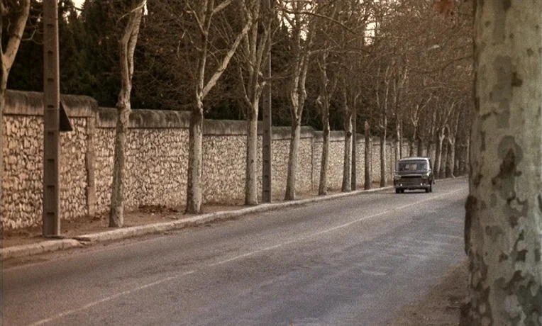 The Man Who Loved Women (1977) movie still: wide — A rural road runs alongside a long stone wall lined with leafless trees. A dark vintage car…