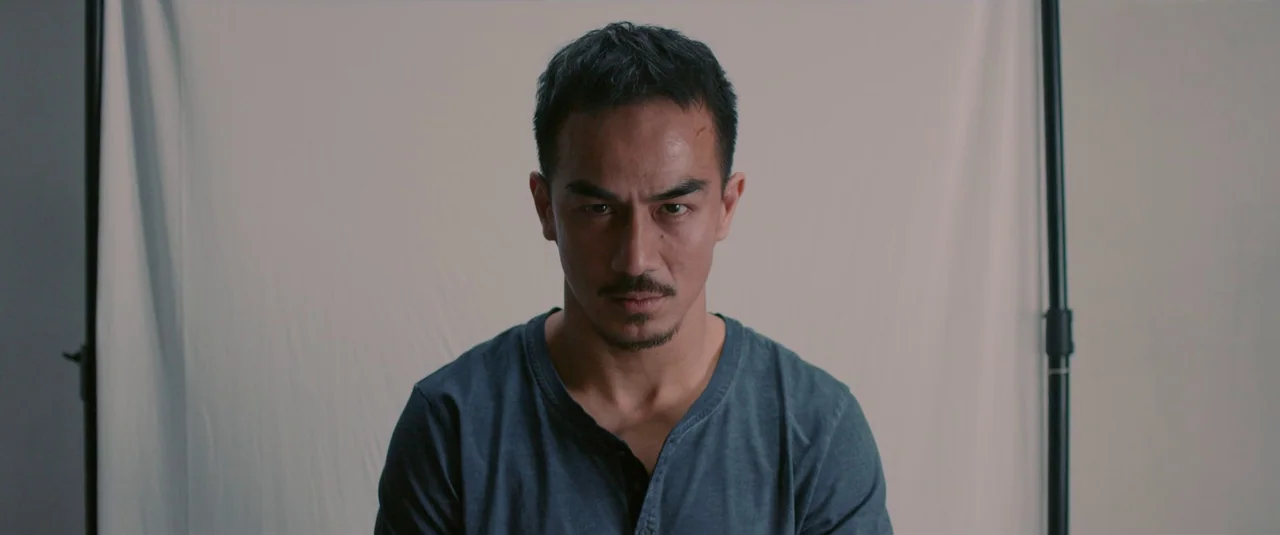 The Night Comes for Us (2018) movie still: medium — A man stands in front of a white photographic backdrop in a studio. The subject looks directly…
