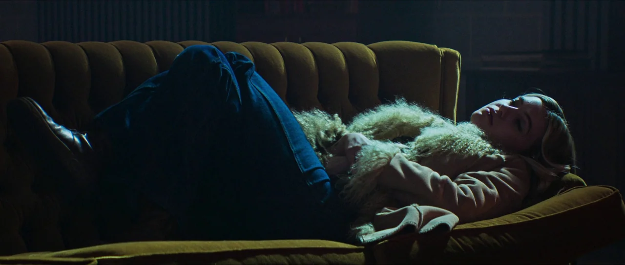 The Sacrifice Game (2023) movie still: medium — A dark, dimly lit room features a velvet-textured mustard yellow couch. A woman with blonde hair…