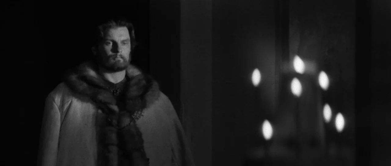 Andrei Rublev (1966) movie still: medium — A stone interior space features a man dressed in historical attire. He stands motionless as a candelabra…
