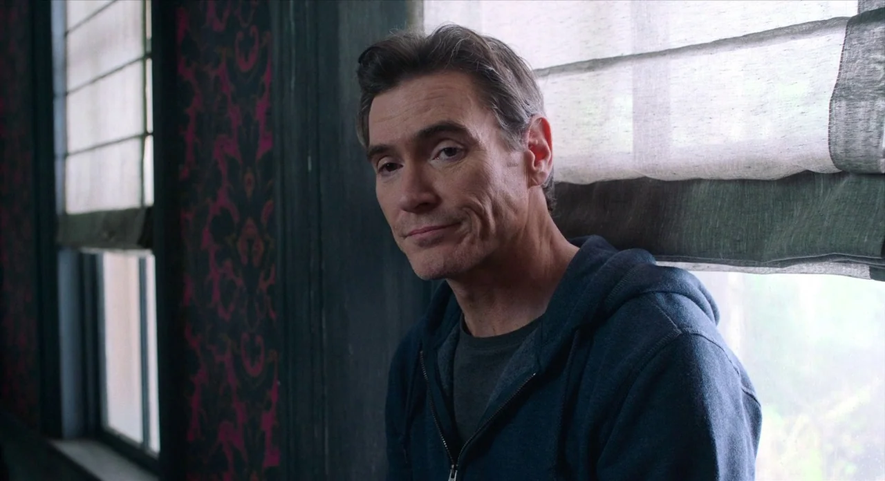 Where’d You Go Bernadette (2019) movie still: medium — A man stands indoors next to a window with patterned wallpaper in the background. The man…