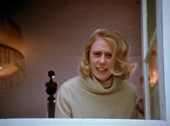 Desperate Living (1977) movie still: medium — A room with a wooden bed post visible on the left and a window frame. A blonde woman in a cream…