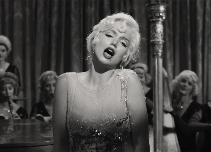 Blonde (2022) movie still: medium — A performance stage with heavy fabric curtains in the background and orchestral musicians present. A woman with…