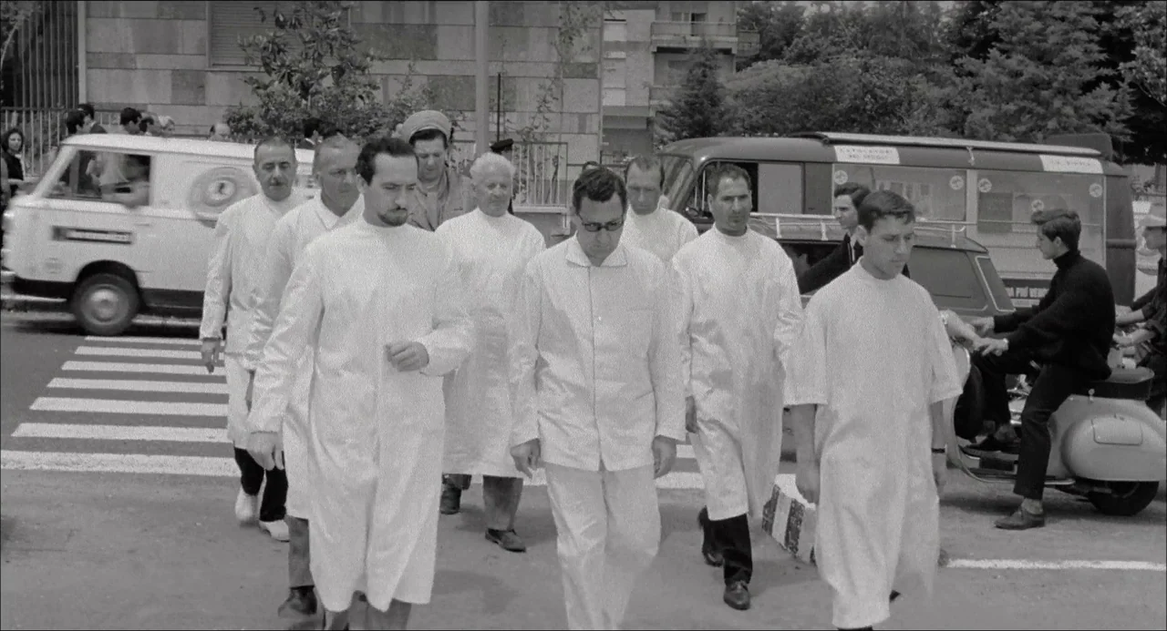 Il Boom (1963) movie still: fullbody — A group of men in white medical coats walk across a city pedestrian crossing. Vehicles are parked along the…