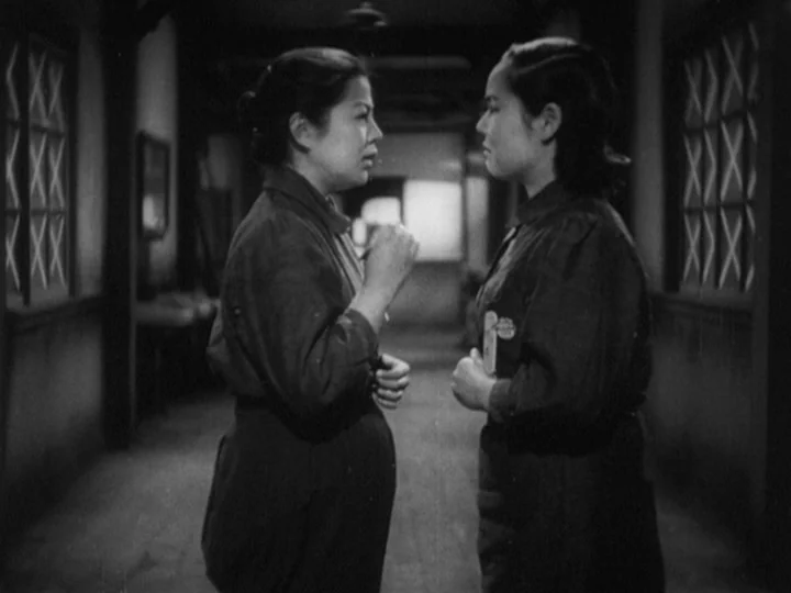 The Most Beautiful (1944) movie still: medium — A dimly lit narrow corridor features lattice-patterned windows on both sides. Two women in work…