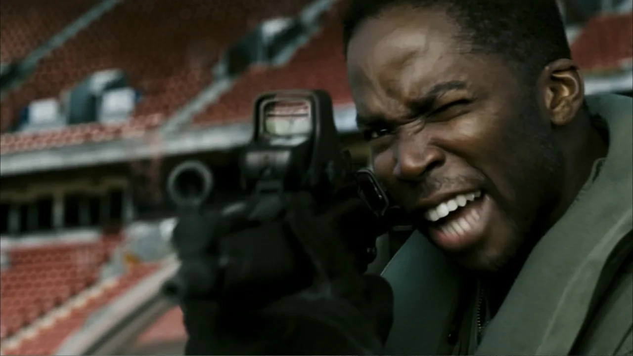 28 Weeks Later (2007) movie still: closeup — A large outdoor stadium has rows of empty red seats in the background. A man in a tactical jacket aims…