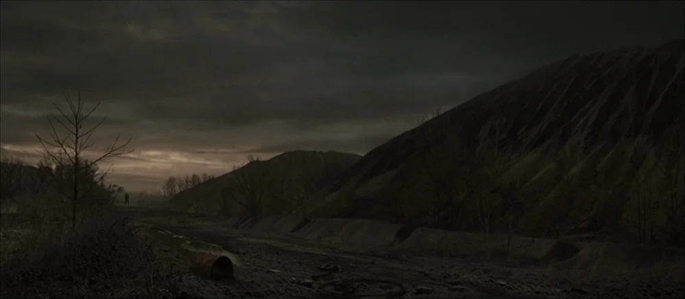 The Road (2009) movie still: wide — A bleak industrial landscape features piles of dark slag and barren, leafless trees. The terrain is composed of…