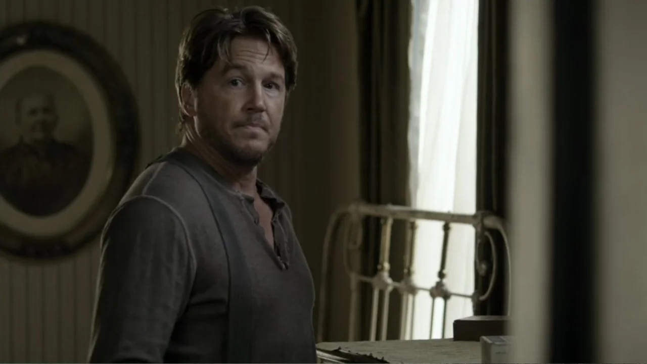 Dawn Rider (2012) movie still: medium — A dimly lit room contains striped wallpaper and a vintage metal bed frame. A man in a grey henley shirt…