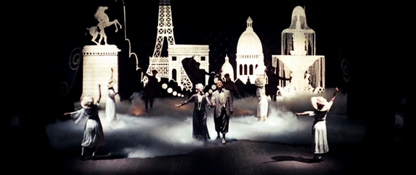 The Boy Friend (1971) movie still: wide — A theatrical stage features large white cutouts of Parisian monuments and a horse. Several performers in…