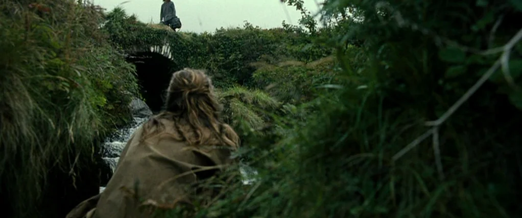 Ondine (2009) movie still: wide — A rural landscape features a small stone bridge covered in thick vegetation. A person crouches in the foreground…