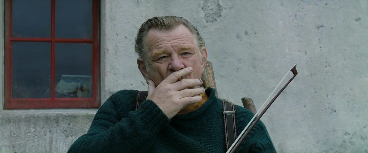 The Banshees of Inisherin (2022) movie still: medium — An older man stands before a weathered white stone wall with a red-framed window. He holds a…