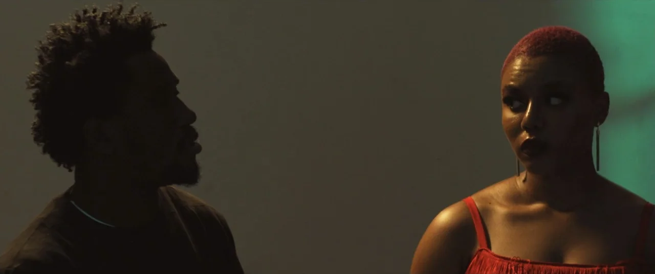 Saving Onome (2024) movie still: medium — A dark interior space with a neutral gray wall and a teal light source. A man and a woman stand facing…