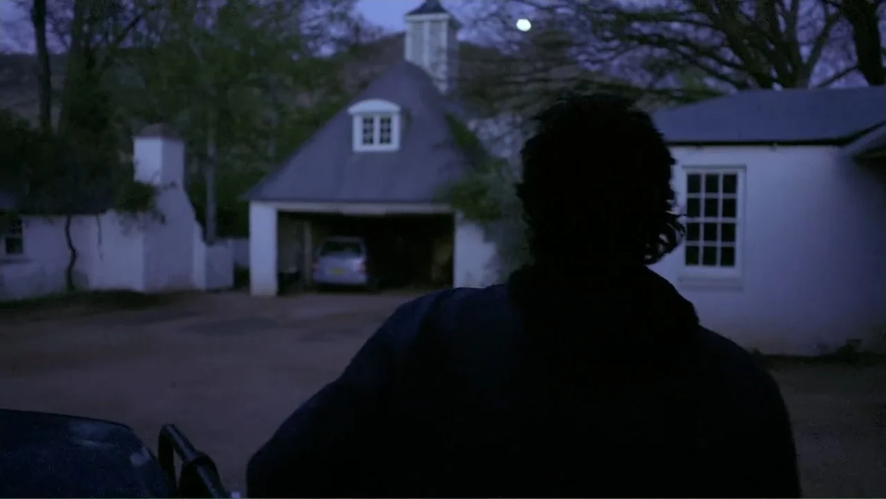 Emily (1976) movie still: medium — A residential driveway at dusk features a white house with a dark garage opening. A person stands in the…