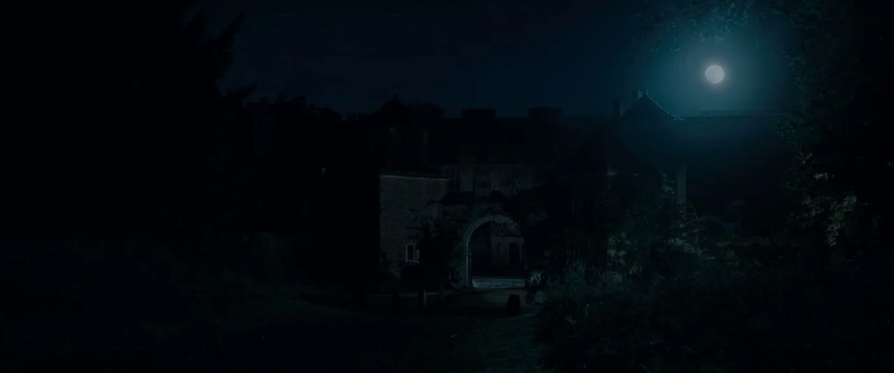 Rebecca movie still: establishing — A dark manor estate architecture is visible under the glow of a full moon. The grounds are filled with dense…