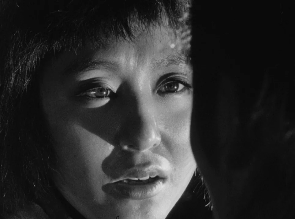 Seven Samurai (1954) movie still: closeup — A dark indoor setting where the face is partially obscured by shadows. The subject gazes towards an…