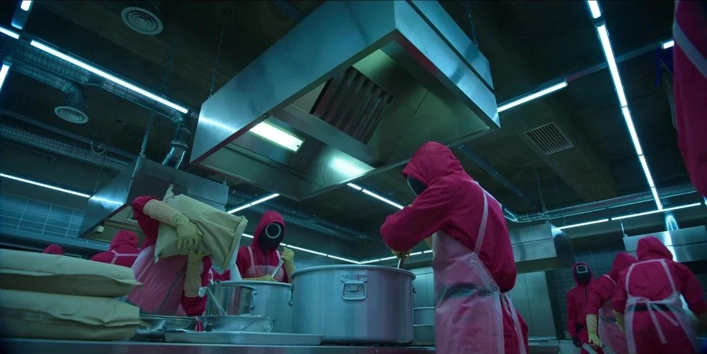 Making of: Squid Game (2021) movie still: wide — An industrial-style food preparation facility features stainless steel hoods and ceiling-mounted…