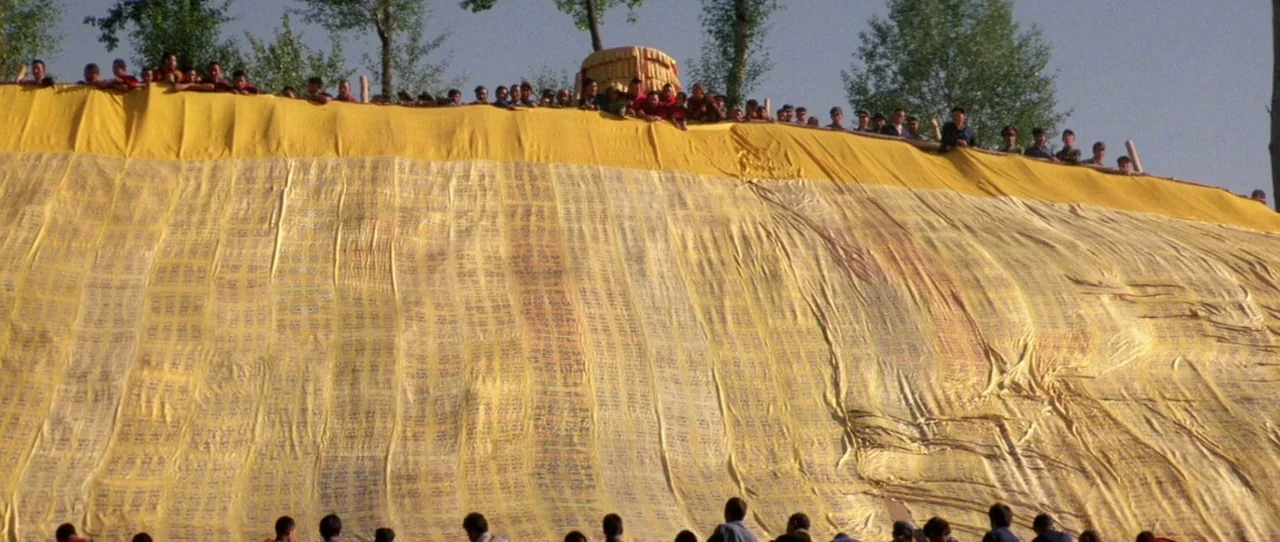 Horse Thief movie still: wide — A massive yellow fabric banner is spread across a sloped outdoor hillside area. People gather along the top edge of…