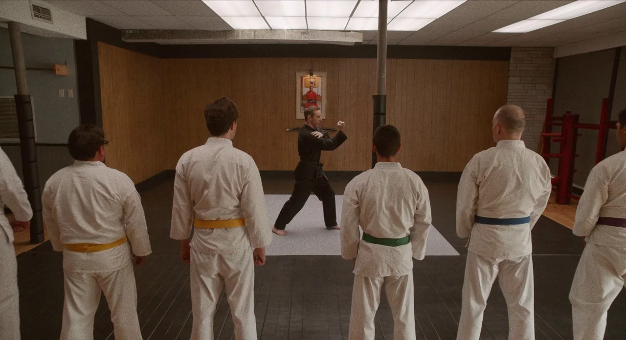 The Art of Self Defence (2019) movie still: wide — A basement studio features wood-paneled walls and a drop ceiling with fluorescent lights. Five…
