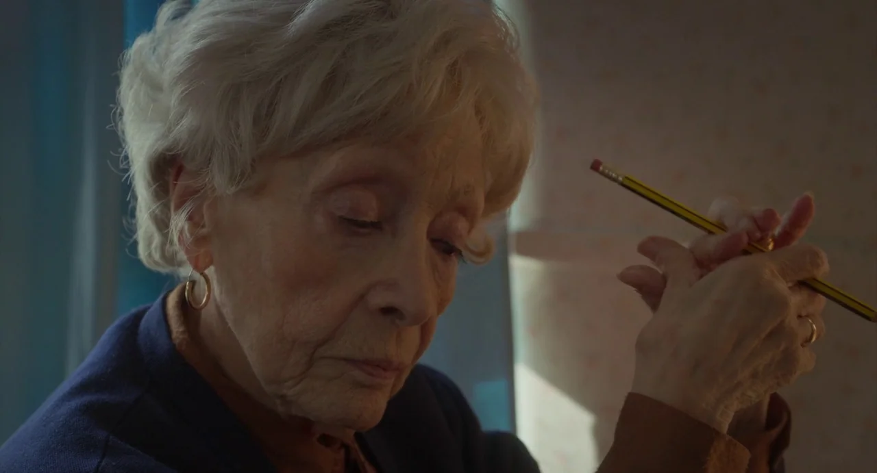 Petite Maman (2021) movie still: closeup — An elderly woman sits indoors with a patterned wallpaper backdrop. She holds a yellow pencil in her hands…