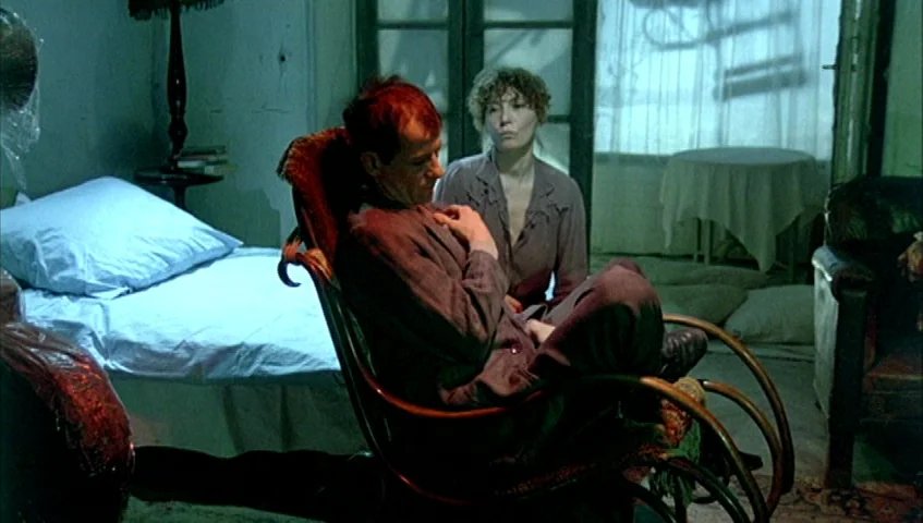 Almanac of Fall (1984) movie still: medium — A dimly lit room contains a bed, a rocking chair, and a draped table. A man and a woman sit in the room…