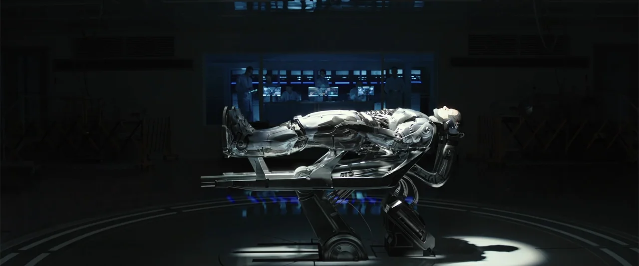 RoboCop (1987) movie still: wide — A dark clinical laboratory features a robotic figure resting on a complex metallic lift. Technicians work in the…