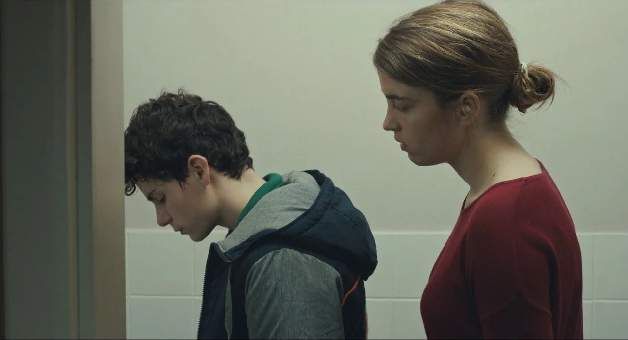 The Unknown Girl (2016) movie still: medium — A sterile, light-colored hallway with a doorframe edge visible on the left side. A teenage boy leans…