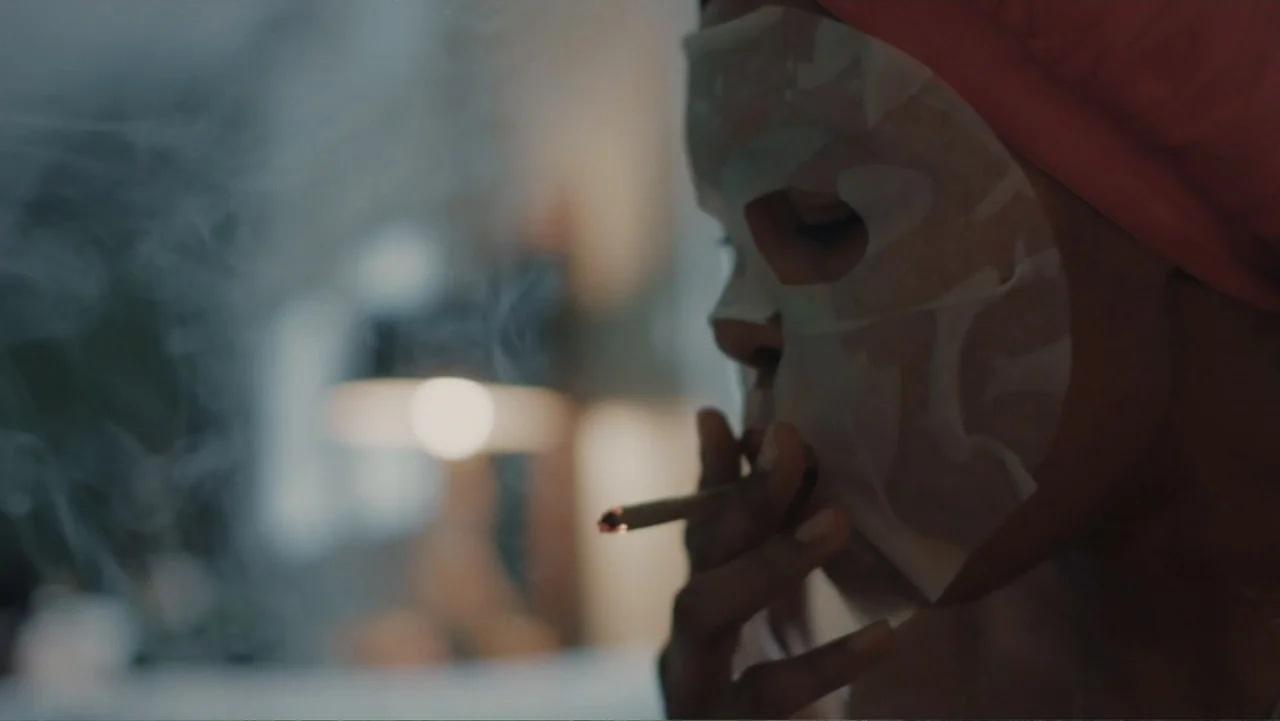 The African Desperate (2022) movie still: closeup — A dimly lit indoor space featuring a person with a white sheet face mask. The subject smokes a…