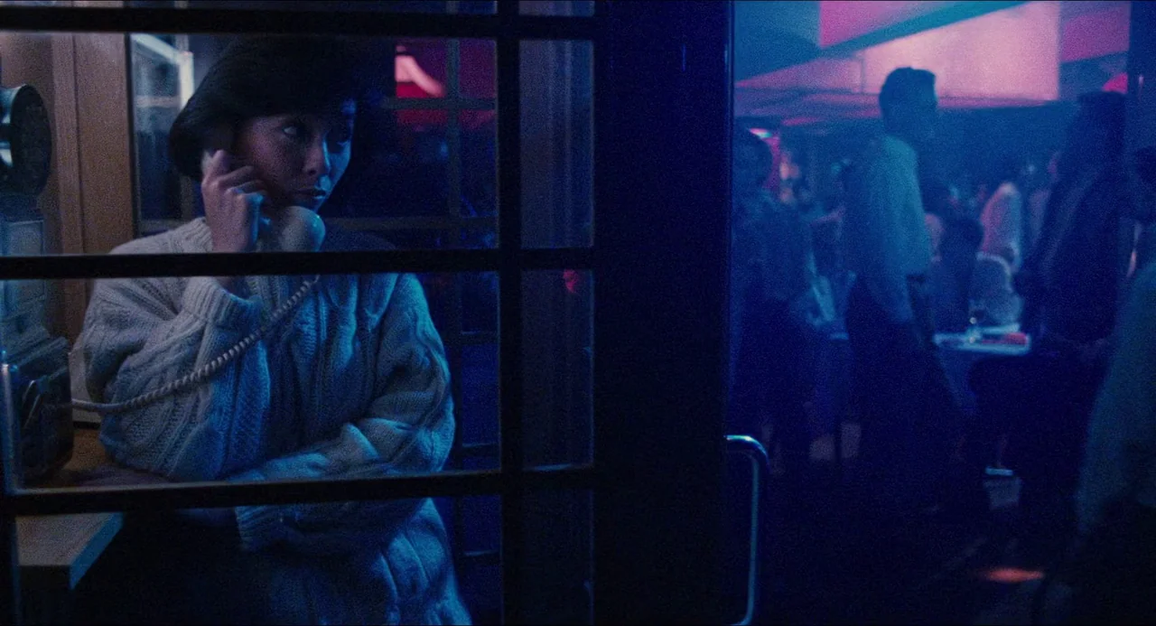 Rouge (1987) movie still: medium — A woman stands inside a glass telephone booth in a dimly lit, blue-hued space. The background shows a blurry bar…