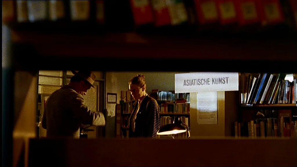 Lola (1981) movie still: wide — A dimly lit room filled with tall shelves of books and paper signs. Two people stand near a desk lamp while browsing…
