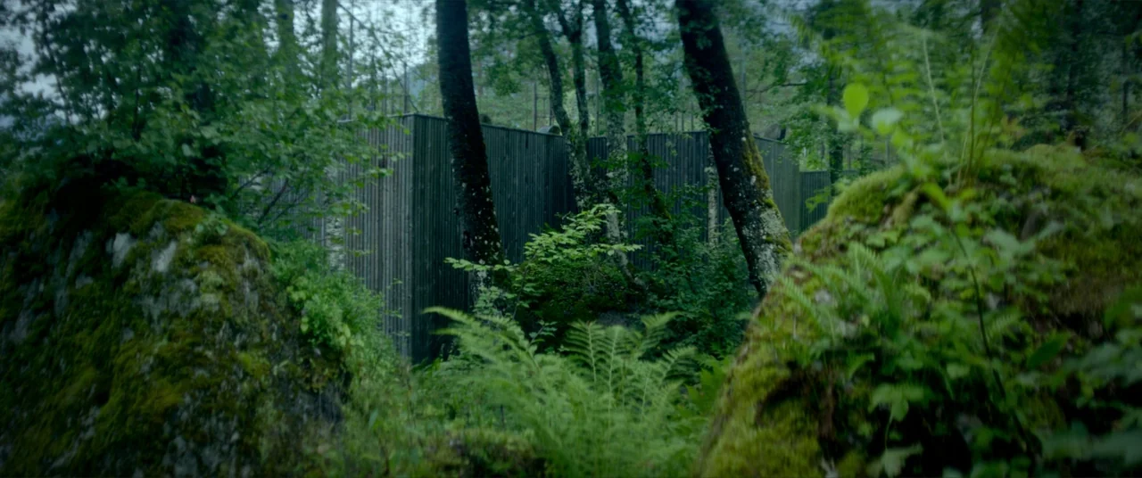 Ex Machina (2015) movie still: establishing — A dark wooden structure with vertical slats is partially obscured by dense overgrown vegetation. Large…