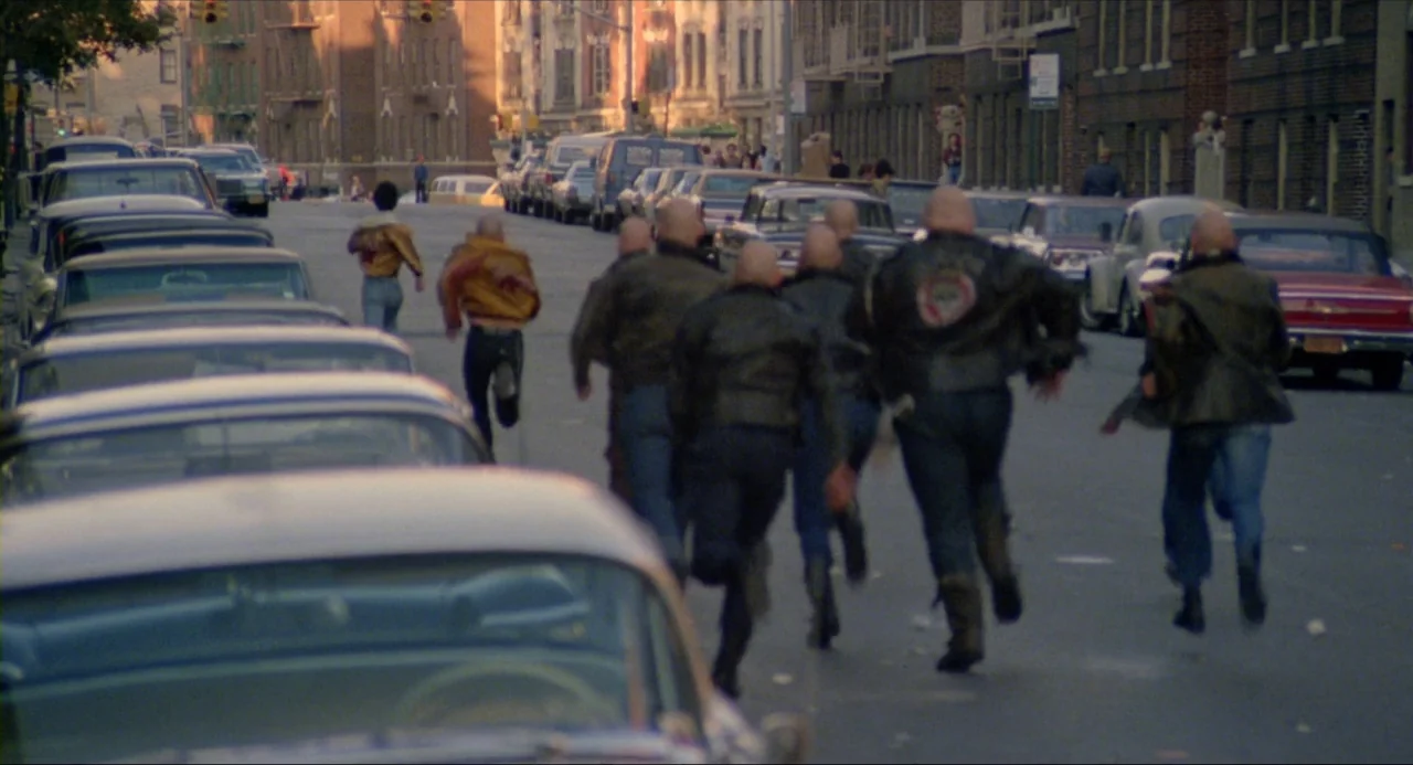 The Wanderers (1979) movie still: wide — A city street lined with parked cars and multi-story brick apartment buildings at dusk. A group of men in…
