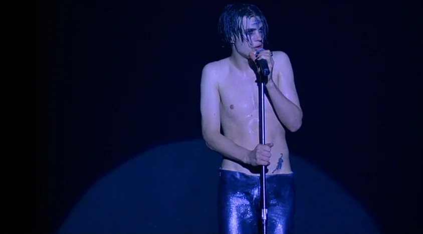 Hedwig and the Angry Inch (2001) movie still: medium — A person stands under a solitary spotlight on a completely dark, featureless stage. The…