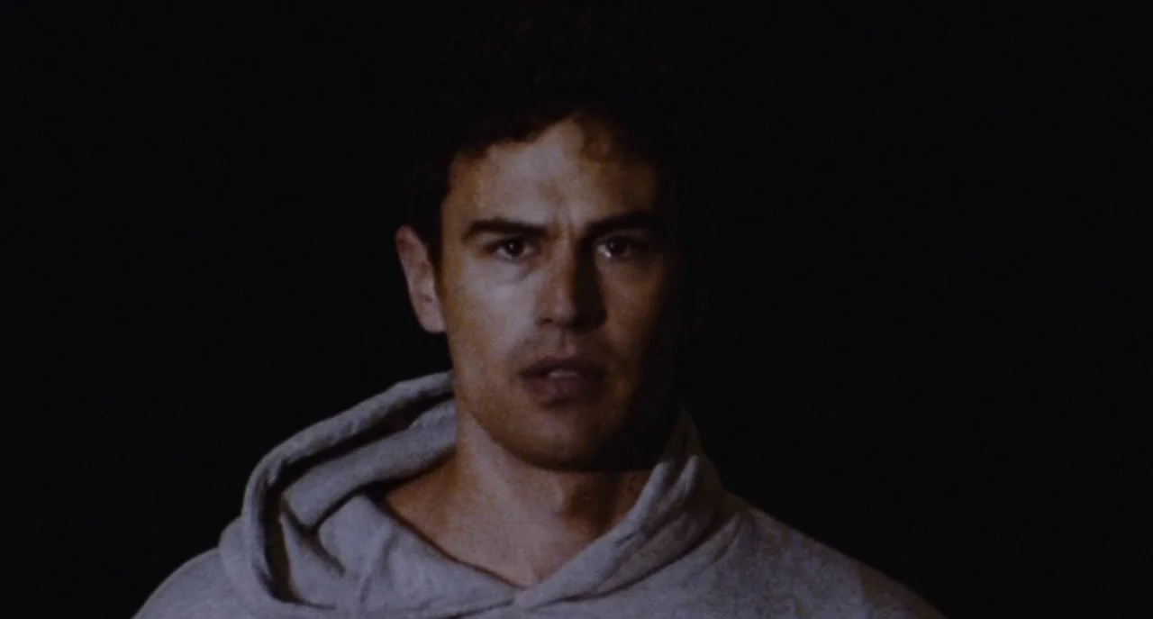 Dual (2022) movie still: closeup — A man stands centered in a completely black, featureless void. He stares forward while wearing a grey hooded…